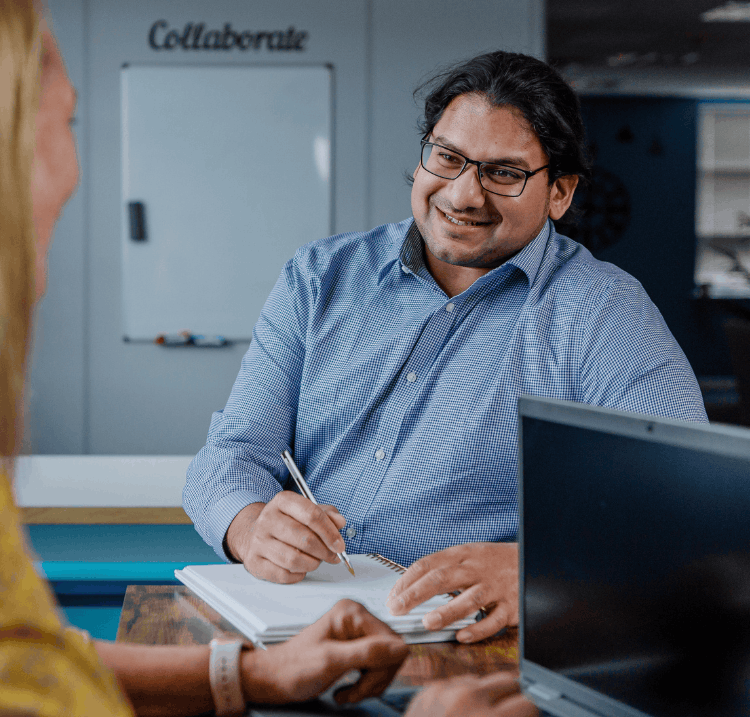 A man with glasses smiling at a woman whilst holding a pen to a notebook