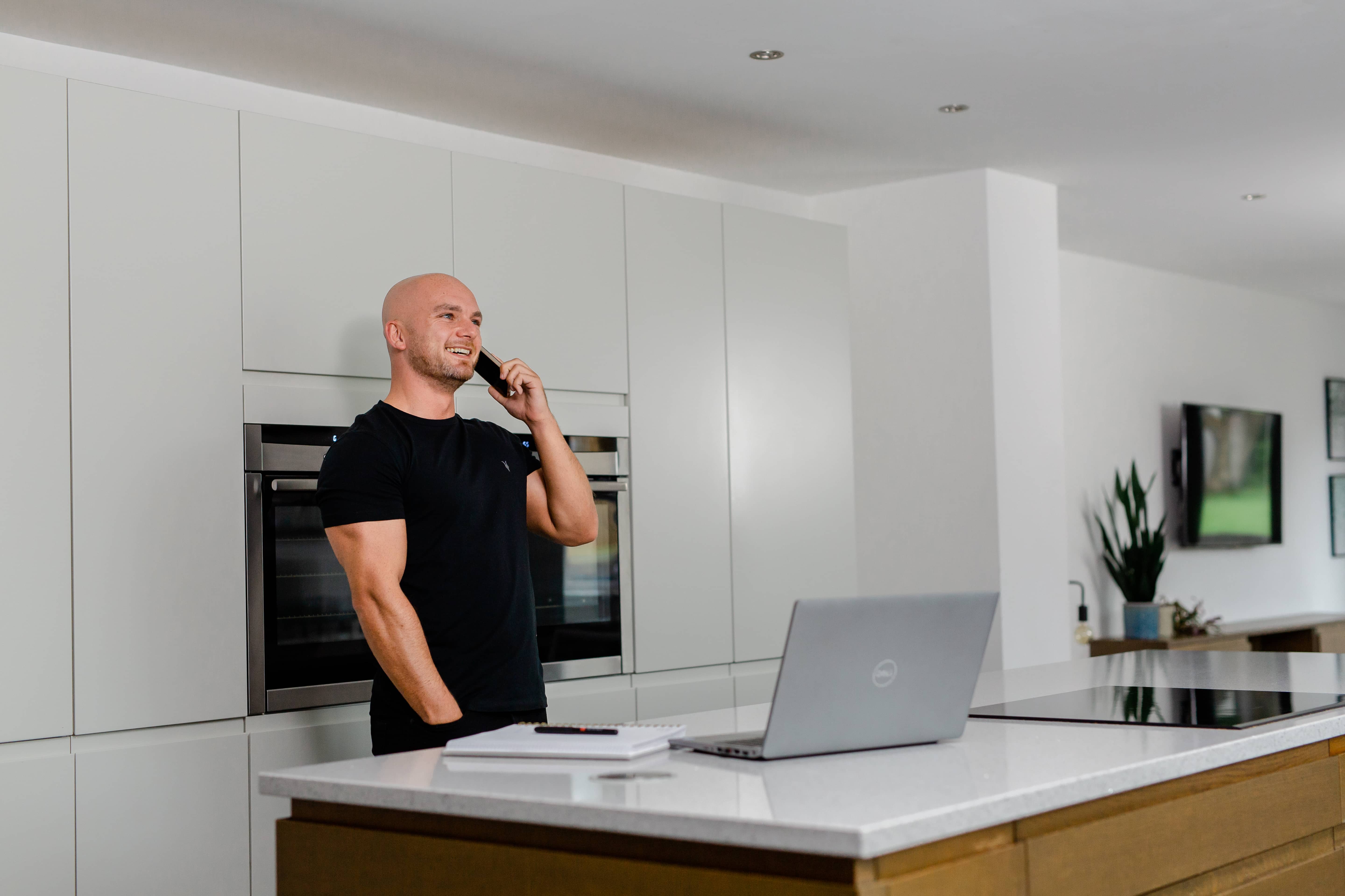 Smiling man in a black t-shirt standing in a modern kitchen with white walls, speaking to a customer on his phone